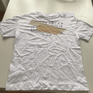 Maison Margirla vit t-shirt med tryck - Vit t-shirt från Maison Margirla med svart tryck fram och bak. Framsidan har diskret logga och bakstycket har ett stort grafiskt tryck med siffror och text. Slim passform och rund halsringning. Passar perfekt som är för dig som är mellan 176-183! Storlek: S. Skick: 10/10! Hör av dig vid intresse eller frågor!