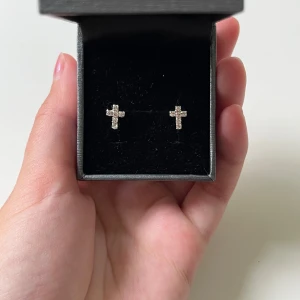 Glittriga korsörhängen i silver - Säljer ett par små örhängen i äkta silver,  i form av kors, täckta med glittrande stenar. I helt nytt skick, De är i silverfärg och har en stilren och modern look som passar perfekt om du gillar detaljer som sticker ut. 