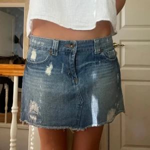 Blå jeanskjol med slitningar - Snygg kort jeanskjol i blå denim med slitna detaljer och rå kant nertill. Klassisk femficksmodell med knapp och dragkedja framtill. Perfekt för en avslappnad och trendig look.