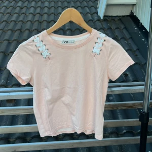 Ljusrosa topp med snörning FB Sister - Supersöt ljusrosa topp från FB Sister i bomull. T-shirten har korta ärmar och snygga vita snörningsdetaljer vid axlarna som ger en unik touch. Perfekt för dig som gillar en enkel men ändå trendig stil.