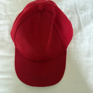 Röd klassisk keps - En stilren röd keps med böjd skärm och sex paneler. Kepsen är tillverkad i ett slitstarkt tyg, perfekt för att lyfta din outfit med en färgklick. Passar både till sportiga och avslappnade looks.