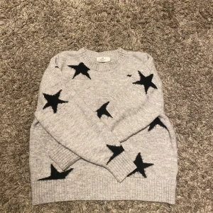Grå stickad tröja med svarta stjärnor H&M - Supermysig grå stickad tröja från H&M med stora svarta stjärnor över hela tröjan. Rund halsringning och långa ärmar med ribbade muddar. Perfekt för dig som gillar en chill och trendig look.