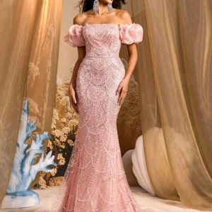 Lång rosa bal- och festklänning - Drömmig långklänning i ljusrosa med glittriga paljetter och pärlor. Offshoulder-modell med puffiga ärmar och snörning i ryggen. Figurnära passform och vackra detaljer som ger en lyxig känsla. Perfekt för bal eller speciella tillfällen.