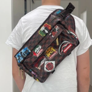 Sprayground brun axelväska limited edition - Limited edition axelväska från Sprayground med brunt rutigt mönster och färgglada patches med olika städer. Väskan har flera fack med dragkedja och bred justerbar rem. Perfekt för dig som vill sticka ut med en unik och streetinspirerad look.