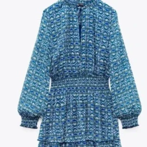 Blå mönstrad kortklänning Zara - Supersöt blå kortklänning från Zara med långa ärmar och smock i midjan. Klänningen har v-ringning och volangdetaljer nertill. Den är oanvänd