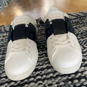 Valentino Garavani Open sneakers vit/blå - Snygga Valentino Garavani Open a kopior sneakers i vitt skinn med bred marinblå rem över mitten. Klassisk rund tå, vita snören och coola nitar på hälen. Perfekt för dig som gillar lyxiga och stilrena sneakers med en twist.  Priset inte hugget i sten