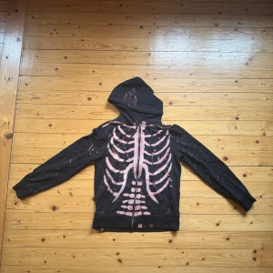 Svart hoodie med skelett och blommor - Cool svart hoodie med dragkedja och huva. Framsidan har ett stort skelett-tryck i rosa och detaljer med blommor på huvan. Hemmagjord design med stänk och långärmad passform. Perfekt för dig som gillar edgy och annorlunda stil.