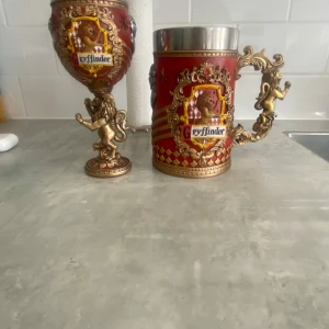 Gryffindor glas  - Bilderna visar två dekorativa dryckeskärl med Gryffindor-tema från Harry Potter, prydda med lejon och detaljer i rött och guld. Dessa är inte böcker utan samlarföremål, perfekta för fans av magi och äventyr. Säljer båda för 900kr om man bara vill ha vin glaset kostar det 300kr och det stora kostar 600kr vin glaset är lite sönder där man håller det så ni vet!
