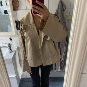 Beige kort trenchcoat från Gina - Säljer en beige kort trenchcoat med bred krage och dubbelknäppning.  Storlek s passar xs och s 
