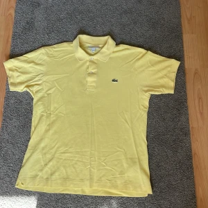 Gul pikétröja från Lacoste, M - Snygg gul pikétröja från Lacoste i klassisk modell med krage och två knappar framtill. Ikoniska krokodilloggan broderad på bröstet. Tröjan är i bomull och har korta ärmar, perfekt för en fräsch och sportig look.