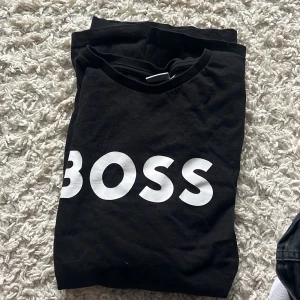 Svart BOSS t-shirt med tryck - Svart t-shirt från BOSS med stort vitt BOSS-tryck på bröstet. Klassisk rund halsringning och korta ärmar. Skön och stilren modell som passar perfekt till jeans eller shorts. Materialet är mjukt och bekvämt, troligtvis bomull.