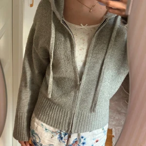Grå hoodie med dragkedja och huva - Mysig grå hoodie med dragkedja och huva. Tröjan har ribbade muddar och snörning vid huvan. Perfekt för lager-på-lager och chill dagar. Passformen är normal och materialet känns stickat och mjukt.