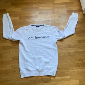 Vit Sail Racing sweatshirt i bomull - Vit sweatshirt från Sail Racing med svart logga på bröstet. Klassisk rund halsringning med diskret broderad detalj i nacken. Tillverkad i mjuk bomull och har långa ärmar samt ribbade muddar för en clean look.