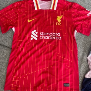 Liverpool matchtröja Nike - Liverpool FCs officiella matchtröja i rött med gula detaljer och tunna gula linjer. Tröjan har klubbmärke och Nike-logga på bröstet samt sponsortryck framtill. Tillverkad i lätt och ventilerande material. Aldrig använd pga fel storlek