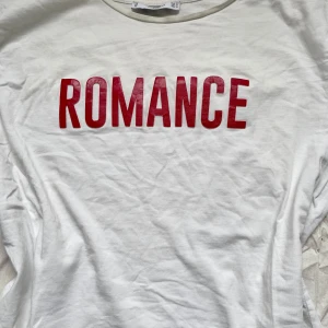 Vit t-shirt med röd text från Mango - Vit t-shirt från Mango Basics med trycket 'ROMANCE' i stora röda bokstäver på bröstet. Klassisk rund halsringning och korta ärmar. Tillverkad i mjuk bomull, perfekt för en chill och stilren look. Väl använd. 