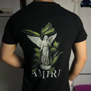 Svart AMIRI t-shirt med ängeltryck - Svart t-shirt från AMIRI med stort tryck av en ängelstaty och gröna blad på ryggen samt AMIRI-logga. Framsidan har loggan med färgstänk som rinner ner. Klassisk passform och rund hals. Perfekt för dig som gillar streetwear med konstnärlig vibe.