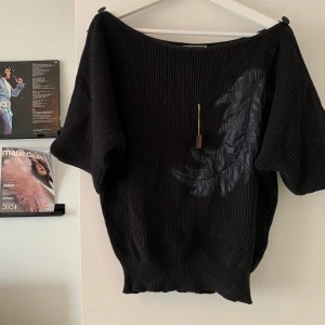 Svart stickad tröja med fjäderdetalj - Svart stickad tröja med bred halsringning och korta ärmar som kan knäppas med knapp. På framsidan finns en stor fjäderdetalj i glansigt tyg. Tröjan har en loose passform och ribbad struktur, perfekt för dig som gillar unika detaljer.