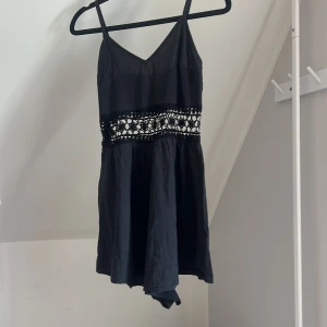 Svart kort byxdress med spetsdetalj - Supersöt svart kort byxdress med smala axelband och v-ringning. Midjan har en bred, genombruten spetsdetalj.  Materialet känns lätt och luftigt. Vet inte storleken men passar Xs kanske S