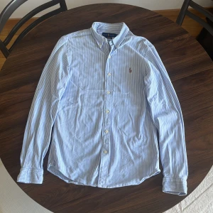 Randig blå skjorta från Ralph Lauren - Snygg blå- och vitrandig skjorta från Ralph Lauren i slim fit-modell. Skjortan har klassisk button down-krage, långa ärmar och broderad logga på bröstet. Tillverkad i mjuk bomull och stängs med vita knappar framtill.
