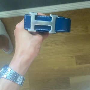 Blått bälte skärp - Säljer ett stilrent blått skärp med ikoniskt H-spänne i silverfärgad metall. Bältet har vita kontrastsömmar och är gjort i skinn. Perfekt accessoar för att lyfta din outfit med en lyxig touch.