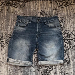 Blå jeansshorts från Jack & Jones - Snygga blå jeansshorts från Jack & Jones i klassisk femficksmodell. Shortsen har uppvikta benslut, normal passform och coola slitningar för en avslappnad vibe. Perfekta för sommardagar när du vill ha en chill och stilren look.