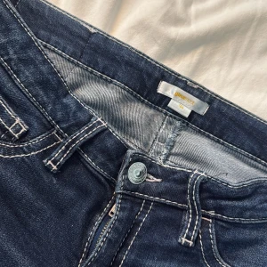 Bootcut jeans från Gina Tricot, mörkblå - Mörkblå bootcut jeans från Gina Tricot med kontrastsömmar och snygga fickdetaljer bak med knappar. Klassisk femficksmodell i stretchig denim som sitter skönt och har en markerad midja. Perfekta för dig som gillar en retroinspirerad look.