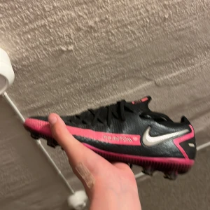 Nike Phantom svart/rosa fotbollsskor - Snygga Nike Phantom fotbollsskor i svart med rosa detaljer och silverfärgad swoosh. Skorna har snörning och mönstrad ovandel i syntetmaterial för extra grepp. Yttersulan har fasta dobbar för bra fäste på planen. Perfekta för dig som vill sticka ut på matchen.