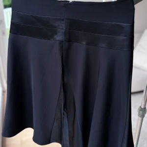 Svart kjol med satindetaljer från Kappahl - Svart, utsvängd kjol från Kappahl med snygga satindetaljer längs midjan och i panelerna fram och bak. Kjolen har en stilren look och är tillverkad i ett mjukt, lätt material som ger ett fint fall. Perfekt för dig som gillar minimalistisk och elegant stil.