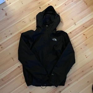 Svart regnjacka The North Face DryVent - Svart regnjacka från The North Face med DryVent-teknologi. Jackan har huva, dragkedja framtill och elastiska muddar. Perfekt för blåsiga dagar och regn, med stilren logga på bröstet och ryggen. Enkel och clean design som funkar till allt.