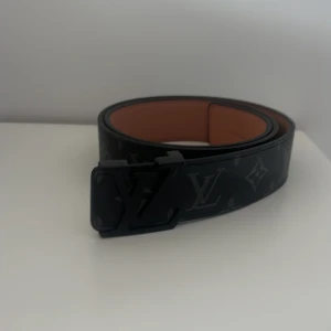 Svart Louis Vuitton bälte i skinn - Snyggt svart bälte från Louis Vuitton med klassiskt monogrammönster och stor LV-spänne framtill. Bältet är gjort i skinn och har en lyxig känsla med mörka detaljer och diskret logga längs hela bandet.