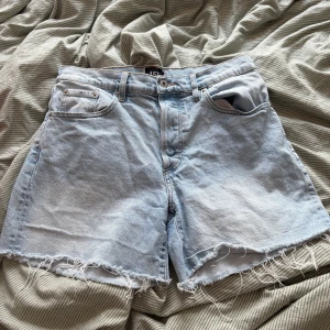 Ljusblå jeansshorts från 157 - Snygga ljusblå jeansshorts från 157 med råa, fransiga kanter och klassisk femficksdesign. Shortsen har normal passform och är perfekta för varma dagar. Tillverkade i jeansmaterial och har bälteshällor samt knapp- och dragkedjestängning.