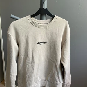 Beige sweatshirt från Jack & Jones - Snygg beige sweatshirt från Jack & Jones Originals Studio med svart tryck på bröstet. Klassisk rund halsringning och långa ärmar. Tillverkad i mjuk bomull, perfekt för en chill och stilren look.