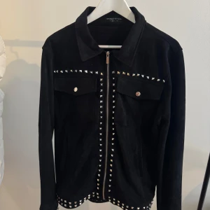 Svart mockajacka med nitar - Svart mockajacka från Manier De Voir med coola silvriga nitar längs bröstet, dragkedjan och nederkanten. Jackan har två bröstfickor med knappar och klassisk krage. Perfekt för dig som vill sticka ut med en cool stil.