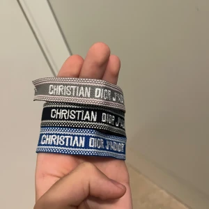 Christian Dior J'Adior armband 3-pack - Tre snygga Christian Dior J'Adior armband i olika färger: grå, svart och blå. Alla är vävda med vit text och har coola tofsar i matchande färger. Perfekt accessoar för att lyfta din outfit med en lyxig touch och street vibe.