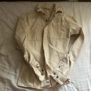 Beige overshirt i bomull från C - Snygg beige overshirt från C i kraftig bomull. Skjortan har två stora bröstfickor, klassisk krage och knappar framtill samt vid ärmsluten. Perfekt att slänga över en t-shirt för en chill look.