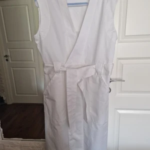 Vit omlottklänning från HOPE - Snygg vit långklänning från HOPE i 100% bomull. Modellen är ärmlös med omlott-design och knytband i midjan för en clean och stilren look. Perfekt för dig som gillar minimalistisk stil och vill ha något bekvämt men ändå trendigt.