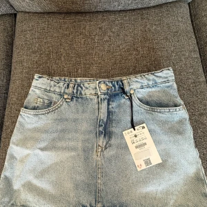 Ljusblå jeanskjol från Bershka - Ljusblå jeanskjol från Bershka med klassisk femficksdesign, bälteshällor och knappstängning framtill. Kjolen har en rak passform och är perfekt för en avslappnad stil. Snygg att matcha med sneakers eller boots.