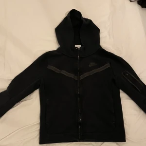 Svart Nike hoodiejacka med dragkedja - Svart hoodiejacka från Nike med hel dragkedja, huva och diskret Nike-logga på bröstet. Jackan har en snygg mörkgrå detalj över bröstet och en praktisk ficka med dragkedja på ena ärmen. Perfekt för en sportig och chill stil. Köptes för 1300kr!. Skriv vid funderingar.