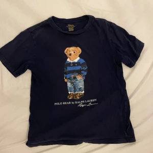 Mörkblå Polo Bear t-shirt Ralph Lauren - Mörkblå t-shirt från Ralph Lauren med ikoniska Polo Bear-trycket framtill. Klassisk passform, rund hals och korta ärmar. Tillverkad i mjuk bomull, perfekt för en chill och stilren look. Nypris 750kr! Skriv vid funderingar🧩