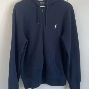 Marinblå hoodie från Polo Ralph Lauren - Snygg marinblå hoodie från Polo Ralph Lauren med dragkedja framtill, huva med snörning och klassisk vit logga på bröstet. Mjuk bomullskvalitet och två fickor framtill. Perfekt för en chill och stilren look. Stl M passar även S, kan även byta med en stl S 