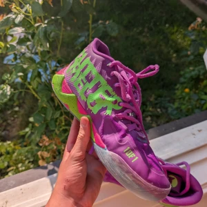 Färgglada sneakers LaMelo Ball x Puma - Unika LaMelo Ball x Puma sneakers i lila och rosa med gröna och blå detaljer. Skorna har coola tryck, snörning och en tecknad katt på sidan. Snygg design med texten 'NOT FROM HERE' och '1 OF 1' på ovandelen. Perfekta för dig som vill sticka ut.