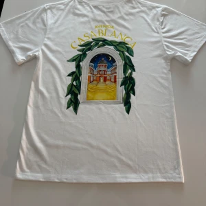Vit Avenida Casablanca t-shirt S - Vit t-shirt i bomull med färgglatt tryck på ryggen som föreställer en fönsterbåge med gröna blad och en byggnad under natthimmel. Texten 'Avenida Casablanca' i gult ovanför motivet. Klassisk passform. Aldrig använt!!