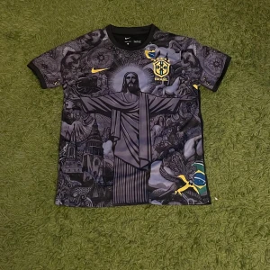 Nike Brasil Jesus - Unik svart Brasilien-fotbollströja från Nike med detaljerad grafisk print av Kristusstatyn och brasilianska symboler. Gula loggor och flaggdetaljer, kortärmad och tillverkad i lätt, sportigt material. Perfekt för dig som vill sticka ut på planen.