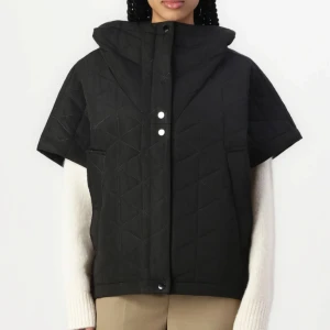 Svart quiltad dunväst med huva - Säljer en svart quiltad dunväst med korta ärmar och stor huva. Västen har tryckknappar framtill och två fickor på sidorna. Snyggt geometriskt mönster i sömmarna ger en modern vibe. Perfekt att slänga över en hoodie eller stickad tröja. Köpt på raglady tara