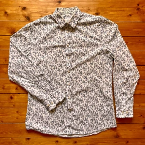 Blommig skjorta från Tiger of Sweden - Snygg vit skjorta från Tiger of Sweden med mörkblått blommigt mönster. Skjortan har klassisk krage, knäppning framtill och långa ärmar. Slim fit-modell i mjuk bomull som känns skön mot huden. Perfekt för dig som vill sticka ut med stilrena detaljer. OBS lite missfärgad kring krage