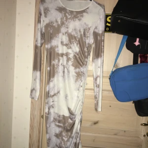 Lång tie-dye klänning med snörning - Superfin långärmad klänning i beige och vitt tie-dye-mönster. Klänningen är ribbad och har en snygg snörningsdetalj längs ena sidan som ger en cool rynkad effekt. Perfekt för dig som gillar unika plagg med lite edge. Tror det är storlek S På sidan kan man göra den mindre genom o dra i snöret 