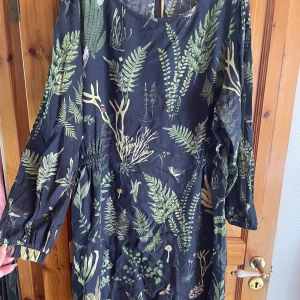 Svart klänning med grönt växtmönster Dedicated - Supersnygg svart klänning från Dedicated med grönt botaniskt mönster av blad och växter. Långärmad modell med avslappnad passform och rund halsringning. Tillverkad i mjuk Tencel lyocell som känns skön mot huden.