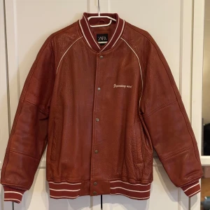 Brun/röd skinn bomberjacka från Zara - Säljer en brun/röd bomberjacka i äkta buffelskinn från Zara. Jackan har vita detaljer, broderad text på bröstet och stor grafisk brodyr på ryggen. Ribbstickade muddar med vita ränder och tryckknappar framtill. Oversized passform. Väldigt fint skick!