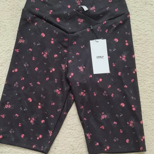 Navy blommiga cykelshorts från ONLY - Svarta cykelshorts från ONLY med små rosa och röda blommor över hela tyget. De har en hög midja med omlott-detalj framtill och är gjorda i ett stretchigt syntetmaterial som sitter skönt på kroppen. Perfekta för en trendig och sportig look.