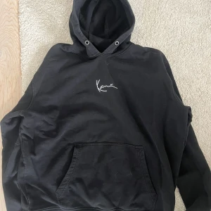 Svart hoodie från Karl Kani - Svart hoodie från Karl Kani med vit broderad logga på bröstet. Klassisk känguruficka framtill och huva med metallöljetter. Skön och avslappnad passform, perfekt till chill dagar. Materialet känns mjukt och är troligtvis bomull eller bomullsblandning.